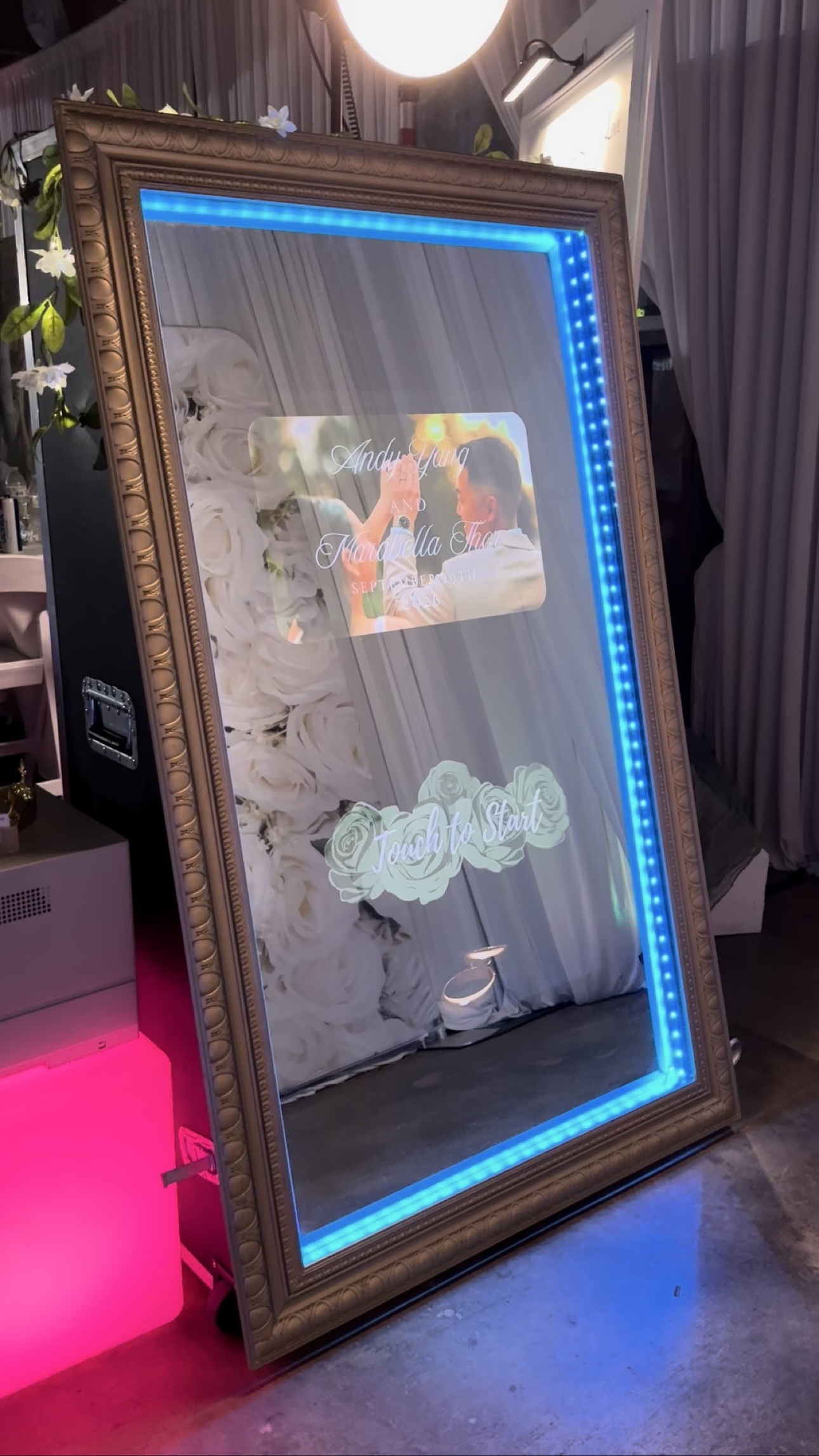 Gold-framed mirror photo booth glowing blue in front of a white flower wall