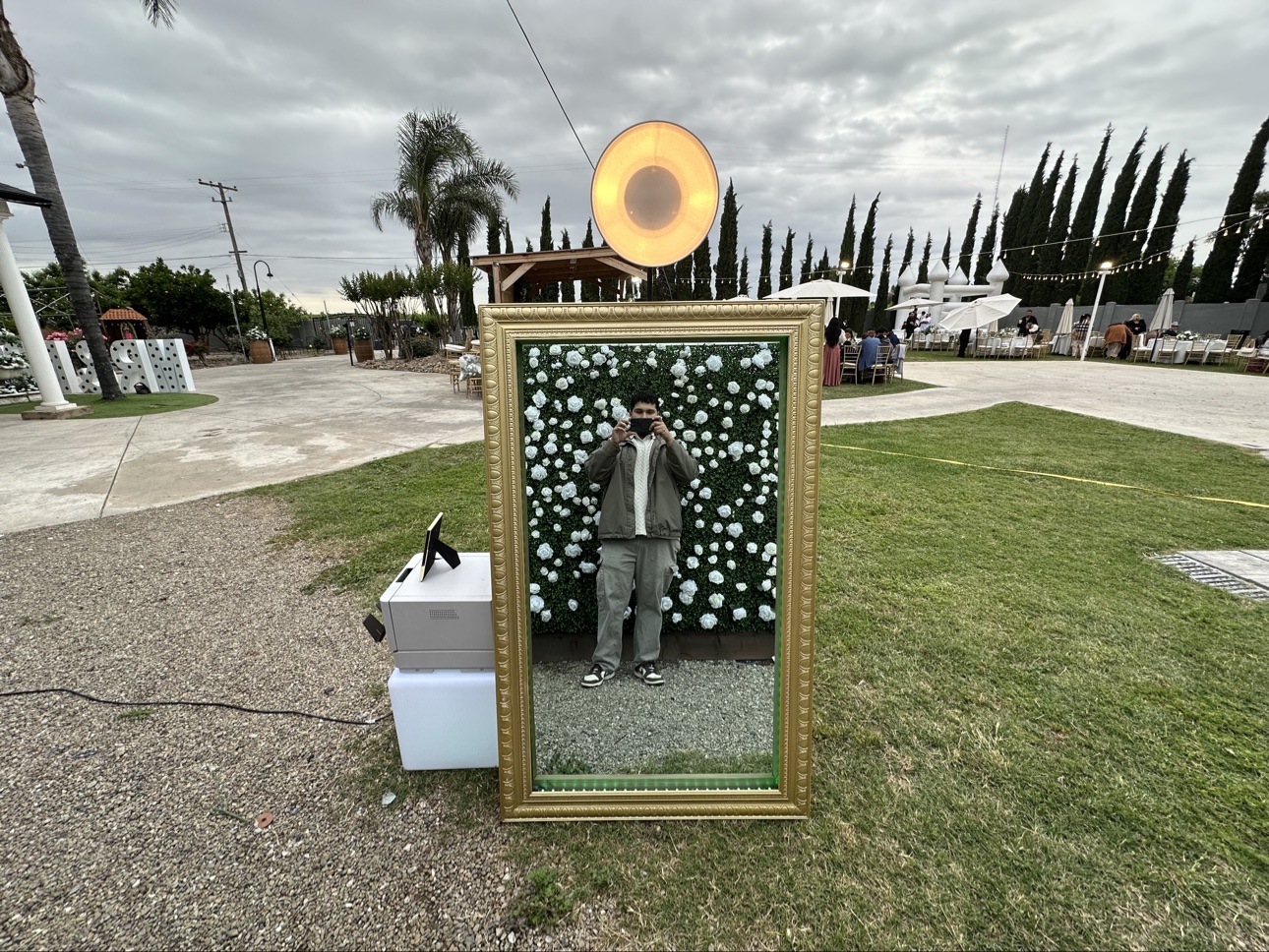 Gold-framed mirror photo booth reflecting a white flower wall at an outdoor celebration