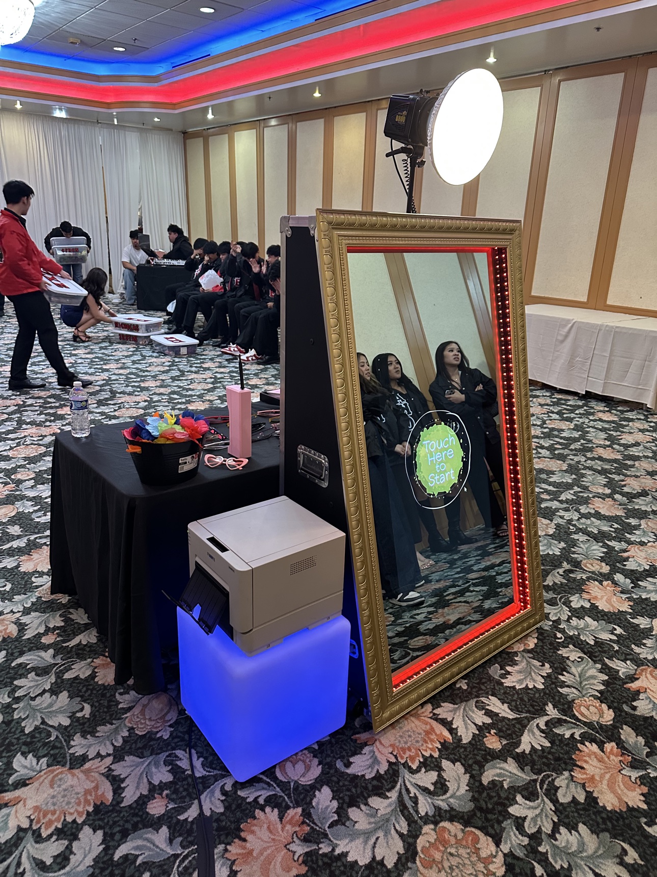 Indoor banquet setup with a red-lit mirror photo booth and a table of colorful props